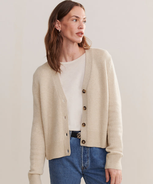 A woman in a relaxed fit Cashmere Colette Cardigan with brown buttons, layered over a white shirt and blue jeans, stands against a plain background, gazing to the side with a neutral expression.