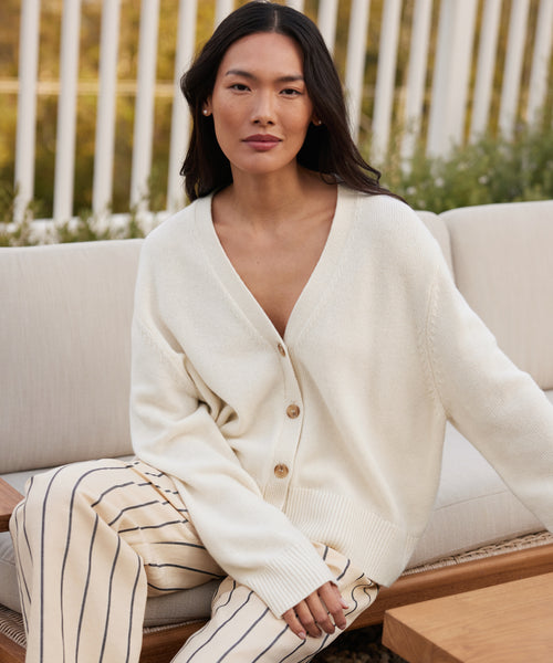 A woman with long dark hair sits on an outdoor sofa, wearing the Cashmere Colette Cardigan and cream pants with black stripes. She gazes calmly at the camera, surrounded by greenery and a white fence in the background.
