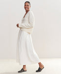 Smiling, a person wears the Cashmere Colette Cardigan made from recycled cashmere, paired with a long white skirt and black sandals, standing against a light neutral background.