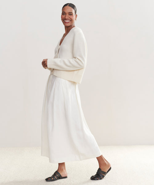 Smiling, a person wears the Cashmere Colette Cardigan made from recycled cashmere, paired with a long white skirt and black sandals, standing against a light neutral background.