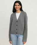 A woman with long dark hair wears the grey Cashmere Colette Cardigan over a white shirt and light blue jeans, standing against a plain, light background and smiling softly.