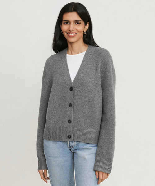 A woman with long dark hair wears the grey Cashmere Colette Cardigan over a white shirt and light blue jeans, standing against a plain, light background and smiling softly.