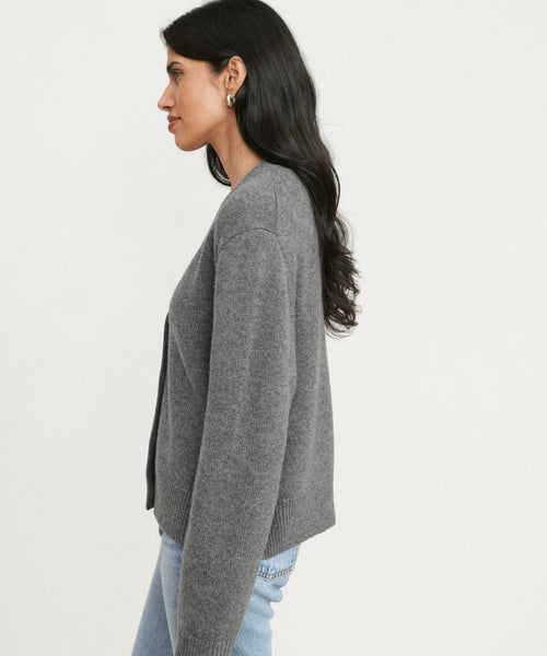 Woman with long dark hair stands in profile against a plain white background, wearing the gray Cashmere Colette Cardigan and light blue jeans.