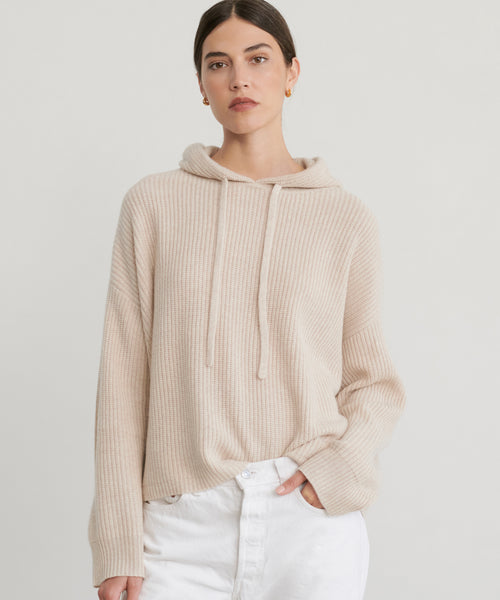 A woman in the Cashmere Fisherman Hoodie, an oatmeal ribbed knit style with drawstrings, pairs it with white pants against a plain light background, one hand in her pocket and a neutral expression.