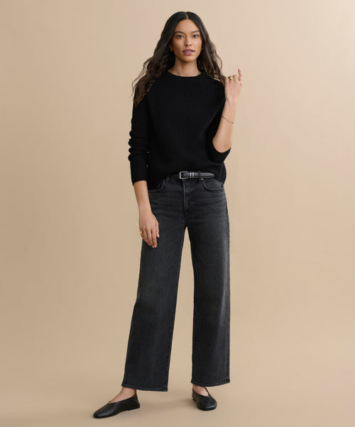 A person with long, wavy hair wears a black Cashmere Fisherman Sweater, black wide-leg jeans, and black flat shoes, standing against a plain beige background.