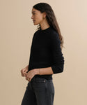 A woman with long wavy hair, wearing the Cashmere Fisherman Sweater in black and dark jeans, stands in profile against a plain beige background.