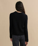 A person with long wavy dark hair is shown from the back, wearing a black Cashmere Fisherman Sweater and dark gray jeans, standing against a plain beige background.