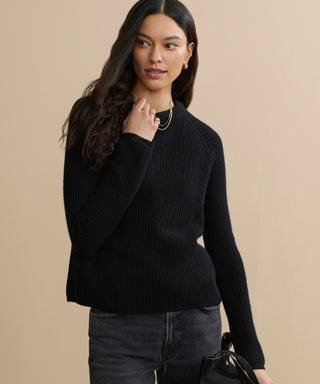 A woman with long, wavy dark hair wears the black Cashmere Fisherman Sweater and dark jeans. She accessorizes with gold earrings, layered necklaces, and holds a black bag while standing against a beige background.