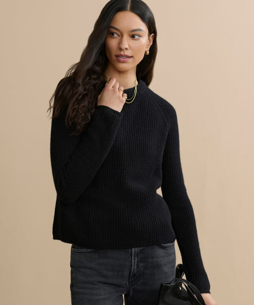 A woman with long, wavy dark hair wears the black Cashmere Fisherman Sweater and dark jeans. She accessorizes with gold earrings, layered necklaces, and holds a black bag while standing against a beige background.