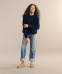 A woman with long curly hair smiles against a beige background, wearing the Cashmere Fisherman Sweater in navy, light blue cuffed jeans, and tan sandals. One arm crosses her body while her other hand rests by her leg.