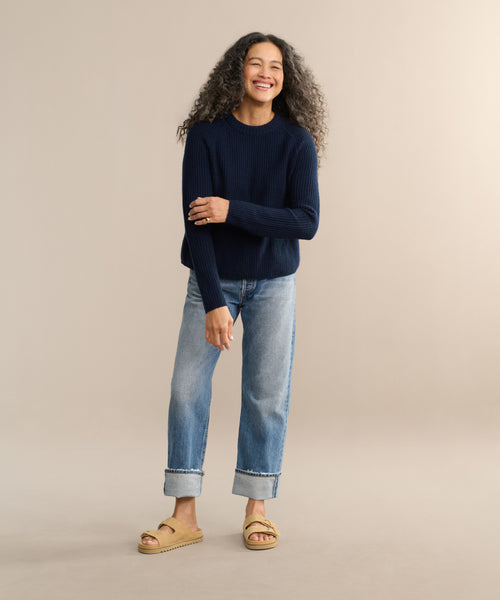 A woman with long curly hair smiles against a beige background, wearing the Cashmere Fisherman Sweater in navy, light blue cuffed jeans, and tan sandals. One arm crosses her body while her other hand rests by her leg.