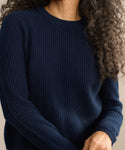A person with long, curly hair models the Cashmere Fisherman Sweater in textured dark blue, shown from shoulders to waist against a neutral background.