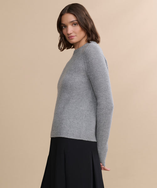 A woman with shoulder-length brown hair models the Cashmere Fisherman Sweater in heather grey with a black skirt, standing sideways against a beige background.