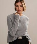 A person with short, light brown hair wears a soft Cashmere Fisherman Sweater in heather grey and black pants with a wavy silver belt buckle, posing against a neutral background.