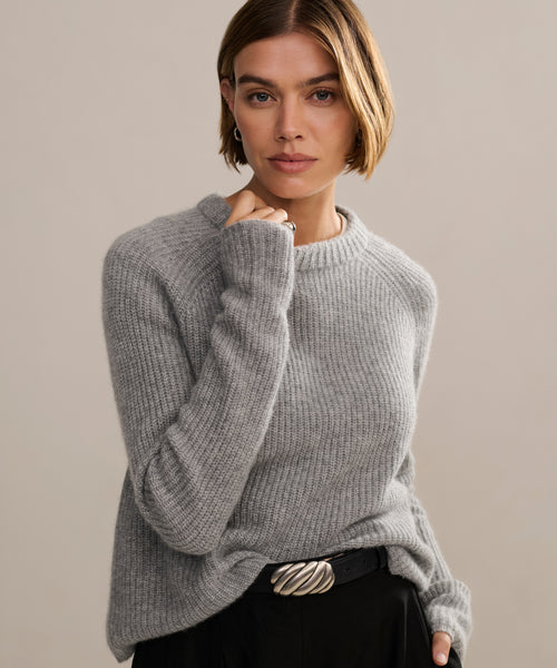 A person with short, light brown hair wears a soft Cashmere Fisherman Sweater in heather grey and black pants with a wavy silver belt buckle, posing against a neutral background.