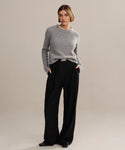 A person with short hair stands confidently against a plain background, wearing a heather grey Cashmere Fisherman Sweater with long sleeves, loose black trousers, and black shoes for a relaxed yet stylish look.