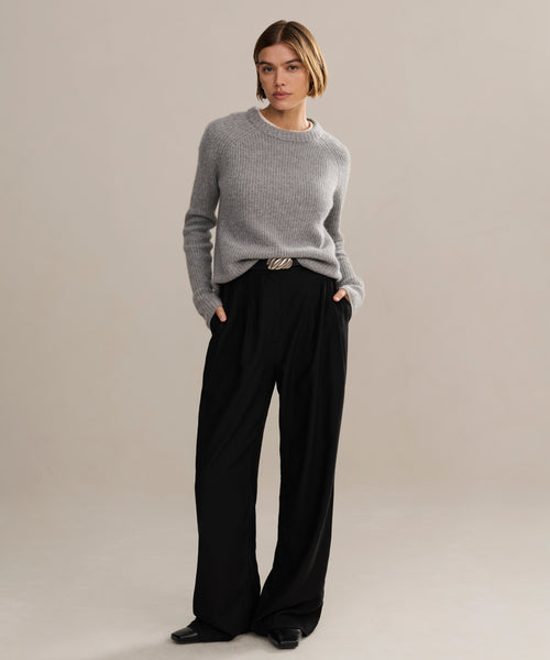 A person with short hair stands confidently against a plain background, wearing a heather grey Cashmere Fisherman Sweater with long sleeves, loose black trousers, and black shoes for a relaxed yet stylish look.