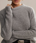 A person wears a cozy Cashmere Fisherman Sweater in heather grey with long sleeves, paired with a black belt featuring a large wavy silver buckle. Only their lower face and upper body are visible.