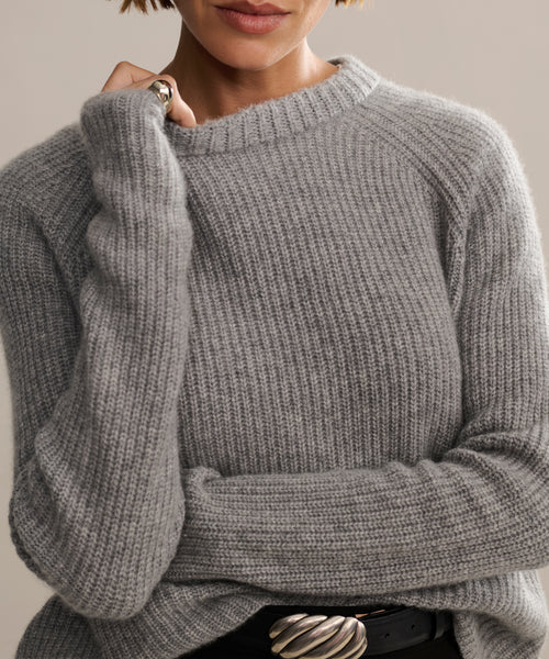 A person wears a cozy Cashmere Fisherman Sweater in heather grey with long sleeves, paired with a black belt featuring a large wavy silver buckle. Only their lower face and upper body are visible.