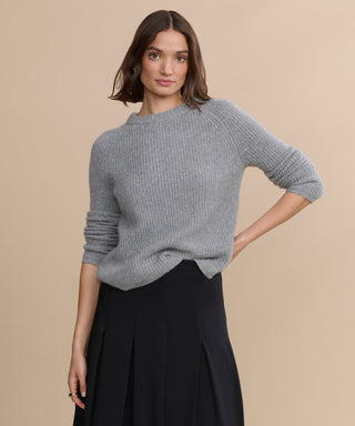 A woman with shoulder-length brown hair wears the Cashmere Fisherman Sweater in heather grey and a black pleated skirt, standing against a plain beige background with one hand on her hip.