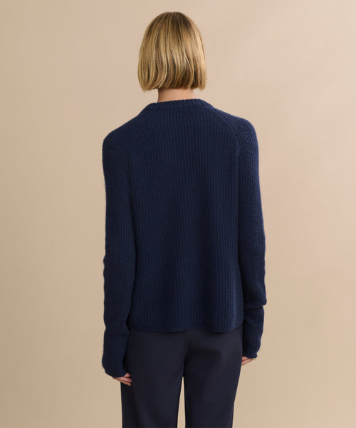 A person with short blonde hair, seen from behind, wears the Cashmere Fisherman Sweater in navy and dark pants, standing against a plain beige background.
