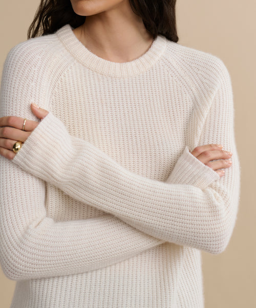 A woman stands with arms crossed, wearing a cozy Cashmere Fisherman Sweater in cream. Gold rings accent her fingers, her face partly out of frame. The background is a soft beige.
