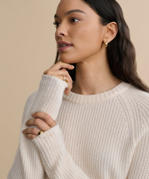 A woman with long dark hair wears a Cashmere Fisherman Sweater in ivory and gold hoop earrings, touching her face thoughtfully to showcase gold rings against a plain beige background.