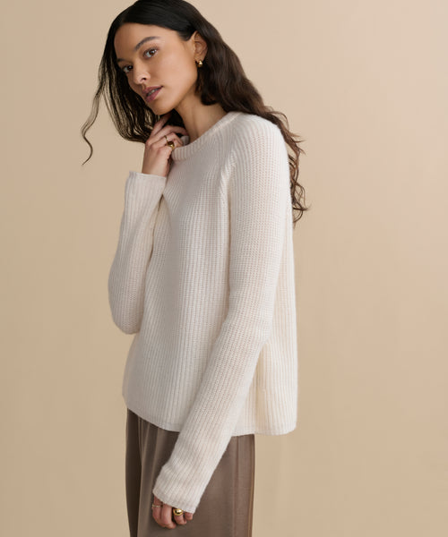 A woman with long, wavy dark hair wears the Cashmere Fisherman Sweater in ivory with brown pants, standing against a plain beige background. She looks slightly to the side, her expression relaxed and thoughtful.