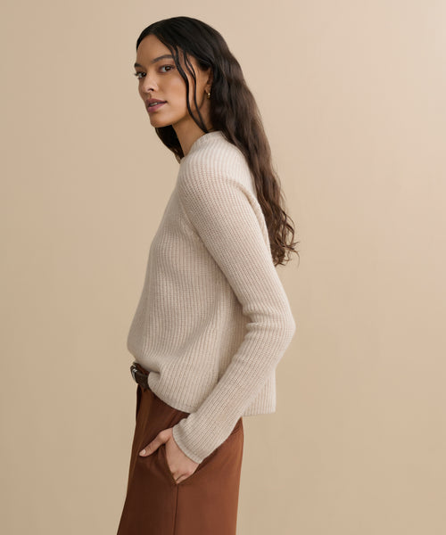 A woman with long, wavy dark hair stands sideways against a beige background, wearing the Cashmere Fisherman Sweater in oatmeal and brown pants, with one hand in her pocket.