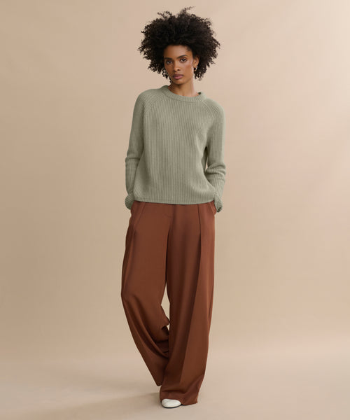 A woman stands against a beige backdrop in a light olive Cashmere Fisherman Sweater and wide brown trousers. With natural curly hair, white shoes, and hands in her pockets, she gazes confidently at the camera.