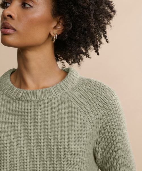 A woman with curly hair and small hoop earrings looks to the side, wearing the Cashmere Fisherman Sweater in light sage green with a textured finish, set against a neutral beige background.