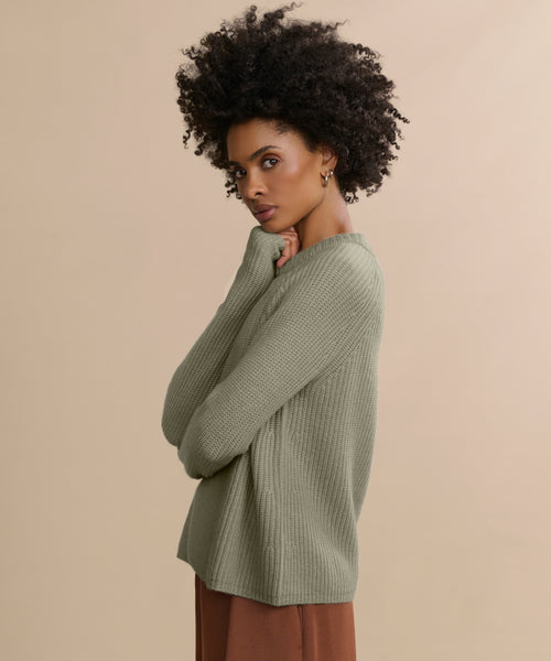Woman with curly hair stands sideways, arms crossed, wearing the Cashmere Fisherman Sweater in light green and brown pants, looking at the camera against a plain beige background.