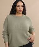A woman with long wavy hair wears the Cashmere Fisherman Sweater in light olive green and black pants, standing against a beige background. She gazes calmly to the side, holding the hem of her sweater.