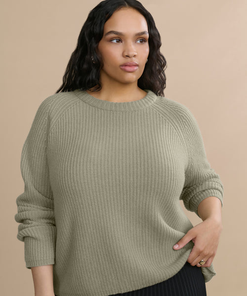 A woman with long wavy hair wears the Cashmere Fisherman Sweater in light olive green and black pants, standing against a beige background. She gazes calmly to the side, holding the hem of her sweater.
