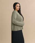 A woman with long, wavy dark hair stands in profile against a beige background, wearing the Cashmere Fisherman Sweater in sage and a long black pleated skirt. She appears relaxed, with her hands resting gently in front of her.