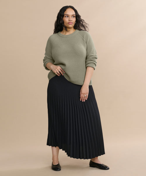 Against a beige background, a woman with wavy dark hair wears the Cashmere Fisherman Sweater in light sage, paired with a black pleated midi skirt and black flats. She has a relaxed, confident expression.