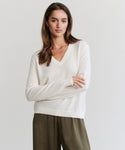 A woman with long brown hair stands with arms crossed against a light background, wearing the Flynn Cashmere Sweater in a relaxed fit cream knit and olive green pants.