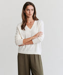 A woman with long brown hair, wearing the Flynn Cashmere Sweater in cream and olive green pants, stands with arms crossed against a plain light background, looking slightly to the side.