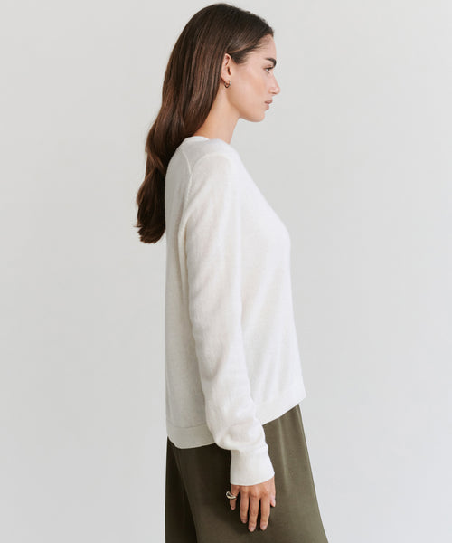 Woman with long brown hair wears the Flynn Cashmere Sweater in white, styled in a relaxed fit, paired with an olive green skirt, standing in profile against a plain light background.
