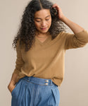 A woman with long, curly hair wears the Flynn Cashmere Sweater in camel, styled with high-waisted blue jeans. She stands with one hand in her pocket and the other touching her hair, looking down with a soft smile.