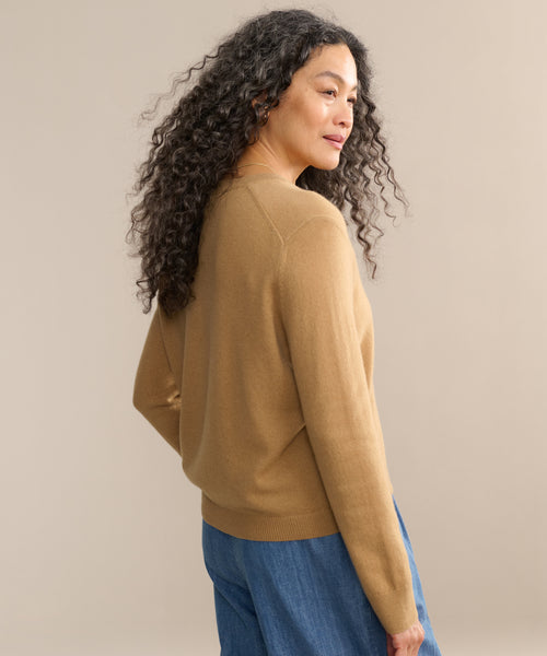 A woman with long curly hair, wearing the Flynn Cashmere Sweater in a relaxed fit and blue pants, stands turned slightly to the side against a plain beige background and smiles gently.