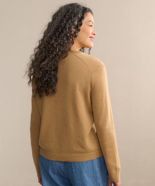 A woman with long, curly hair stands smiling, facing away from the camera. She wears the Flynn Cashmere Sweater in a relaxed fit with blue jeans, set against a plain beige background.