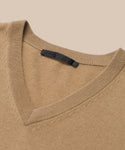 A close-up of the Flynn Cashmere Sweater—a beige V-neck made from soft cashmere, detailed with ribbed edges and a black label inside the collar—set against a neutral beige background.