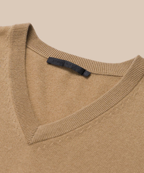 A close-up of the Flynn Cashmere Sweater—a beige V-neck made from soft cashmere, detailed with ribbed edges and a black label inside the collar—set against a neutral beige background.