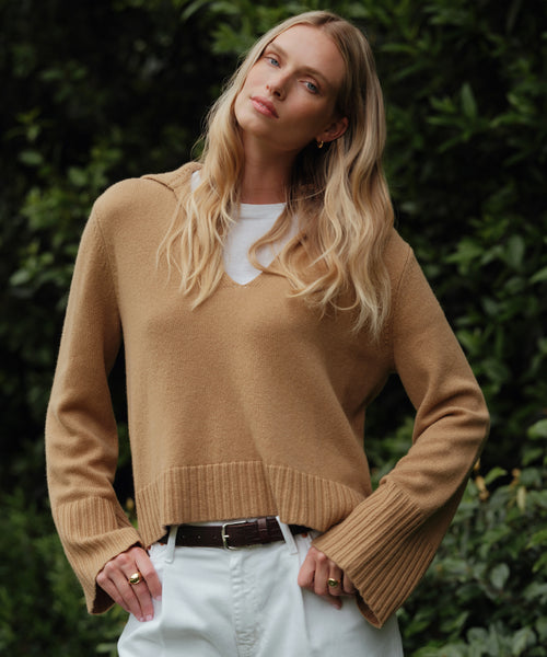 A woman with long blonde hair stands outdoors in front of green foliage, wearing the relaxed fit Cashmere Frances Polo sweater over a white shirt, white pants, and a dark belt, with her hands resting on her hips.