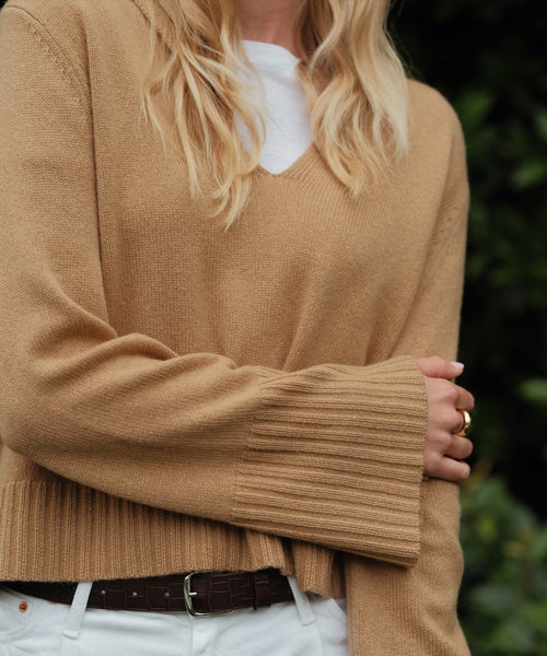 A blonde woman wears the Cashmere Frances Polo, a relaxed-fit camel knit sweater with wide ribbed sleeves, layered over a white shirt and paired with white pants and a brown belt. She stands outdoors with greenery behind her.