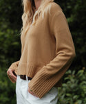 A person with long blonde hair wears the Cashmere Frances Polo, a relaxed fit knit camel-colored sweater with ribbed cuffs, paired with white pants while standing outdoors among greenery.