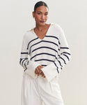 A woman wears the Cashmere Frances Polo—a relaxed fit knit white sweater with bold navy stripes and wide sleeves—paired with white pants. She stands against a plain light background, hair pulled back, looking confidently at the camera.