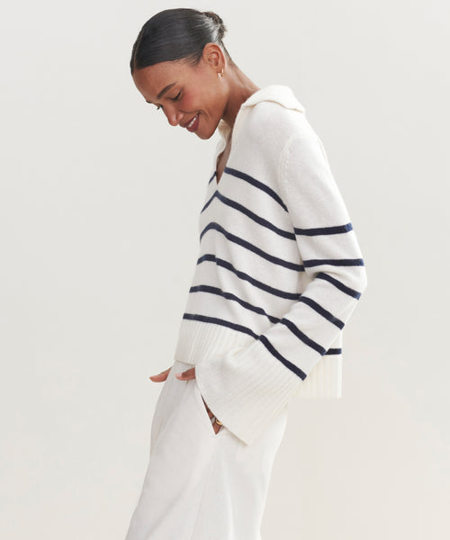 A woman smiles and looks down, wearing the Cashmere Frances Polo with navy stripes and white pants, standing with hands in her pockets against a plain light background.