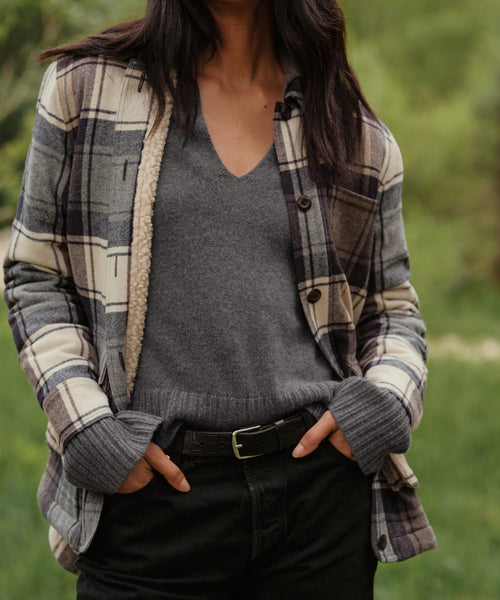 A person stands outdoors in a relaxed fit gray Cashmere Frances Polo, a plaid jacket with sherpa lining, and black pants with a black belt. Their hands are in their pockets, framed by a softly blurred green background.